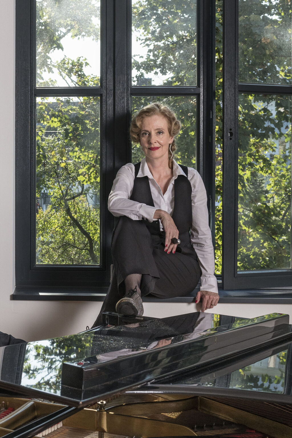 Marie Oser auf der Fensterbank sitzend, die Füße auf dem Flügel. Vor dem Fenster sind grüne Bäume zu sehen