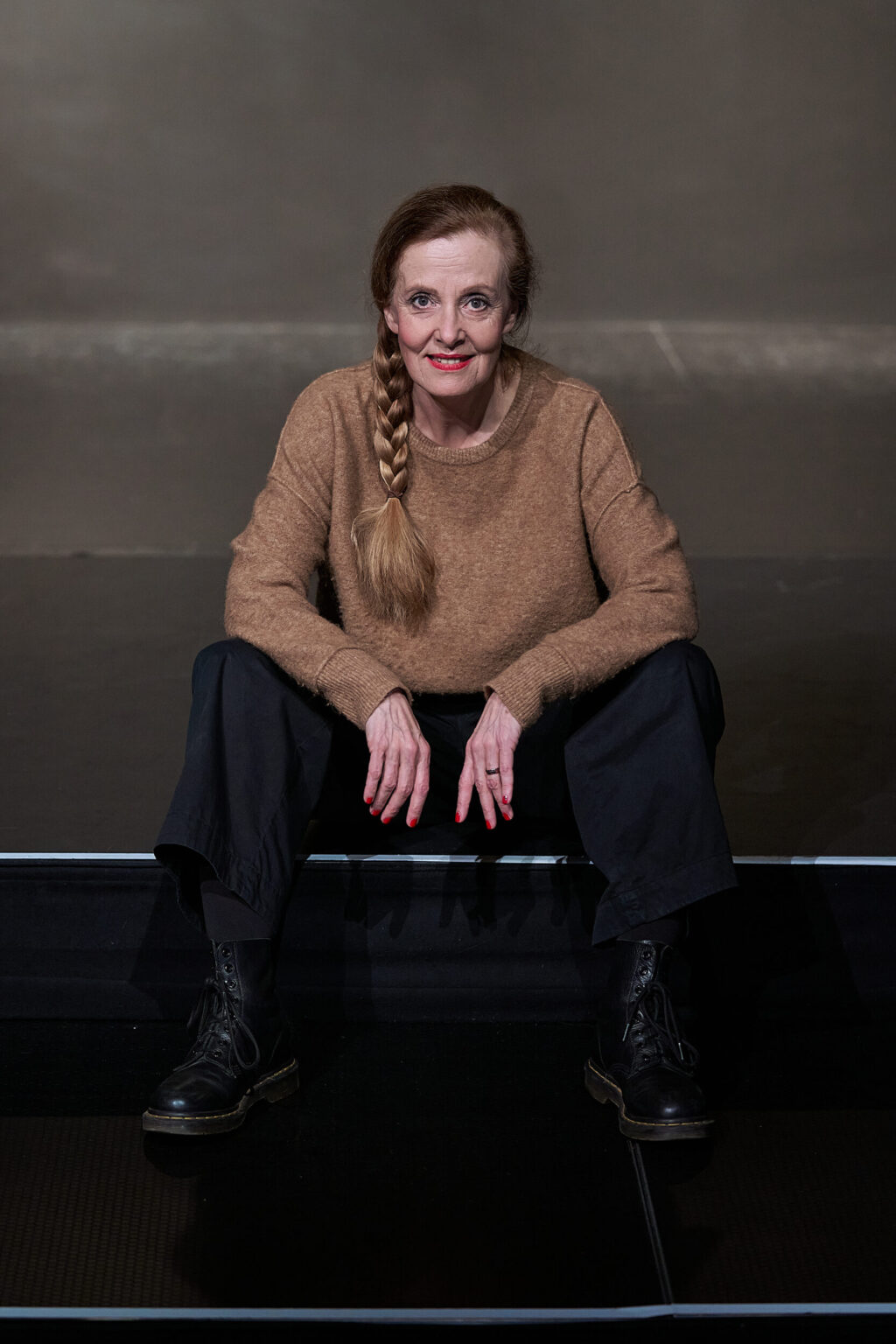 Marie Oser vor grauem Hintergrund auf einer Treppe sitzend, mit Zopf, camelfarbigem Wollpulli, schwarzer Hose und Boots